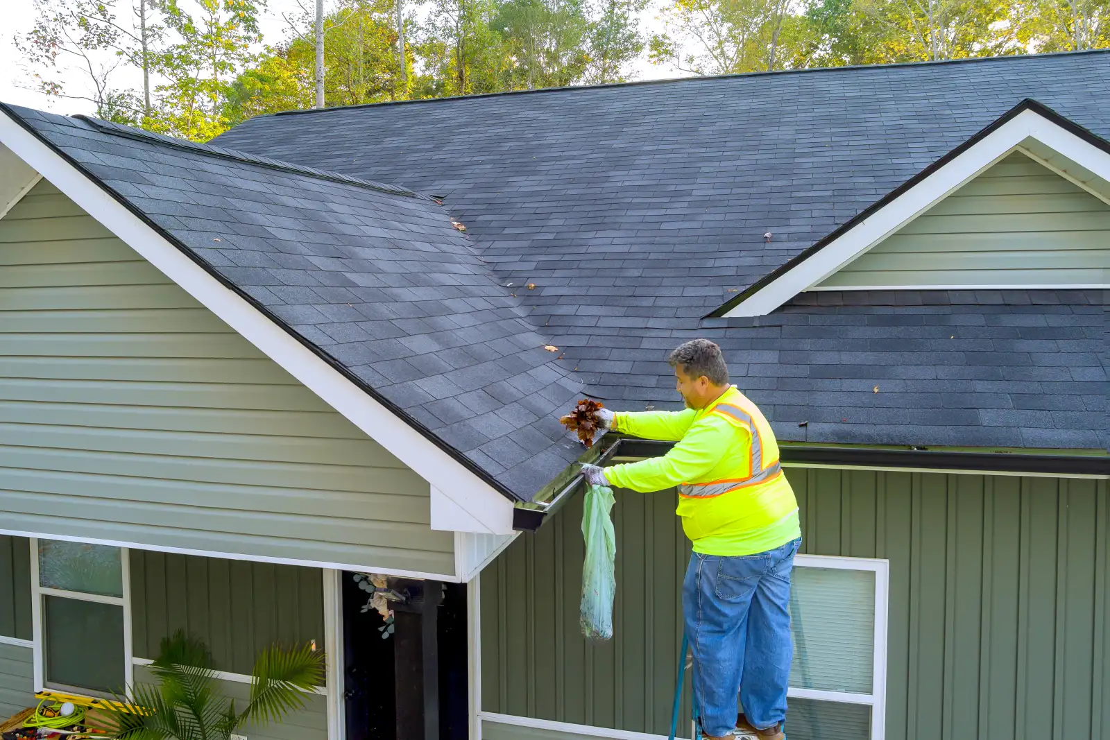 Worker in a safety vest on a ladder pulling wet leaves out of a clogged gutter on a green two story home