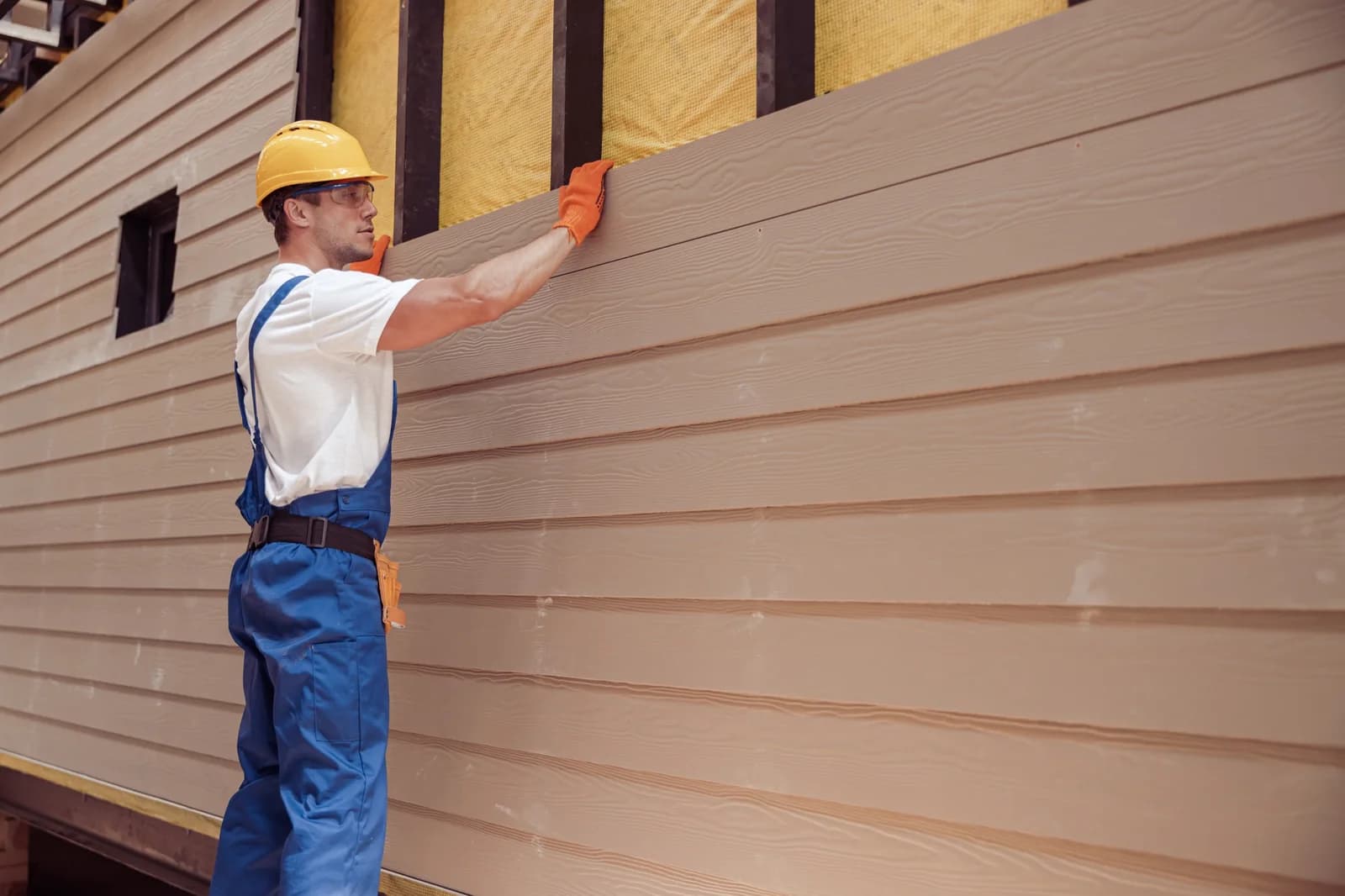 Worker in a hard hat fitting a beige lap siding board onto a house exterior