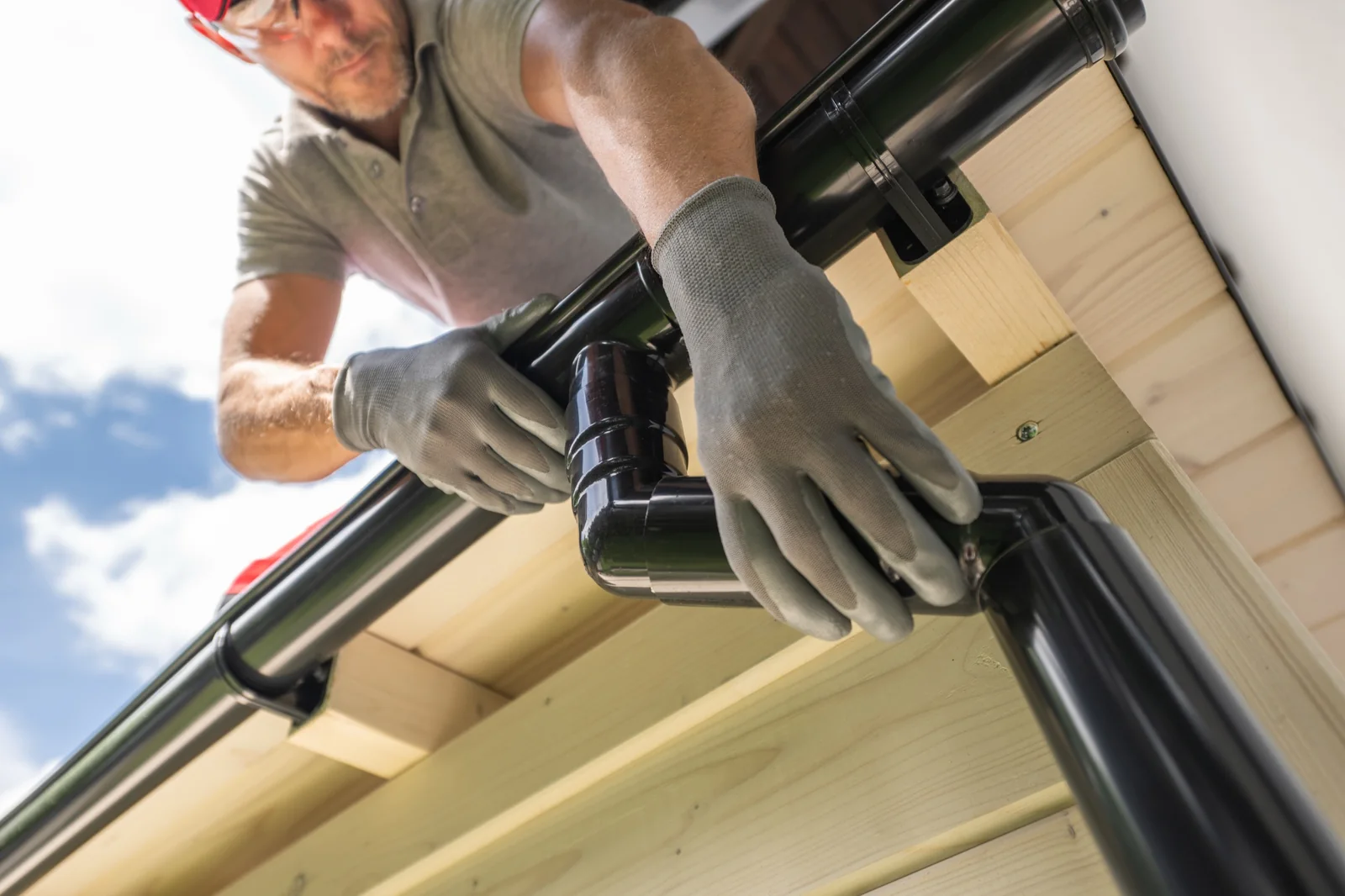 Worker in gloves fitting a black downspout elbow onto a roof gutter